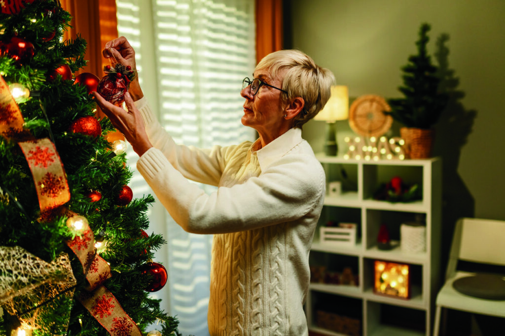 Senior woman is carefully placing a red christmas ornament on a brightly lit tree in her home, adding a touch of festive cheer to the room